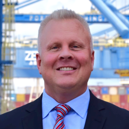 A professional headshot of Hans Bean, Chief Commercial Officer of North Carolina Ports. Container cranes are in the background.